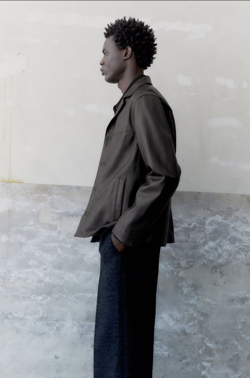 Profile shot of a Black male model in dark jacket and trousers against a textured wall in natural light, fashion editorial.