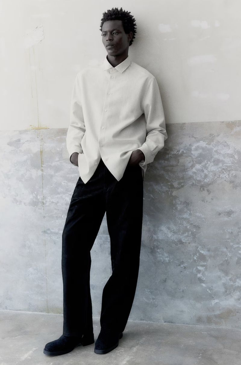 A slender adult male in a white dress shirt and black trousers leans against a textured wall, looking stylish and understated.