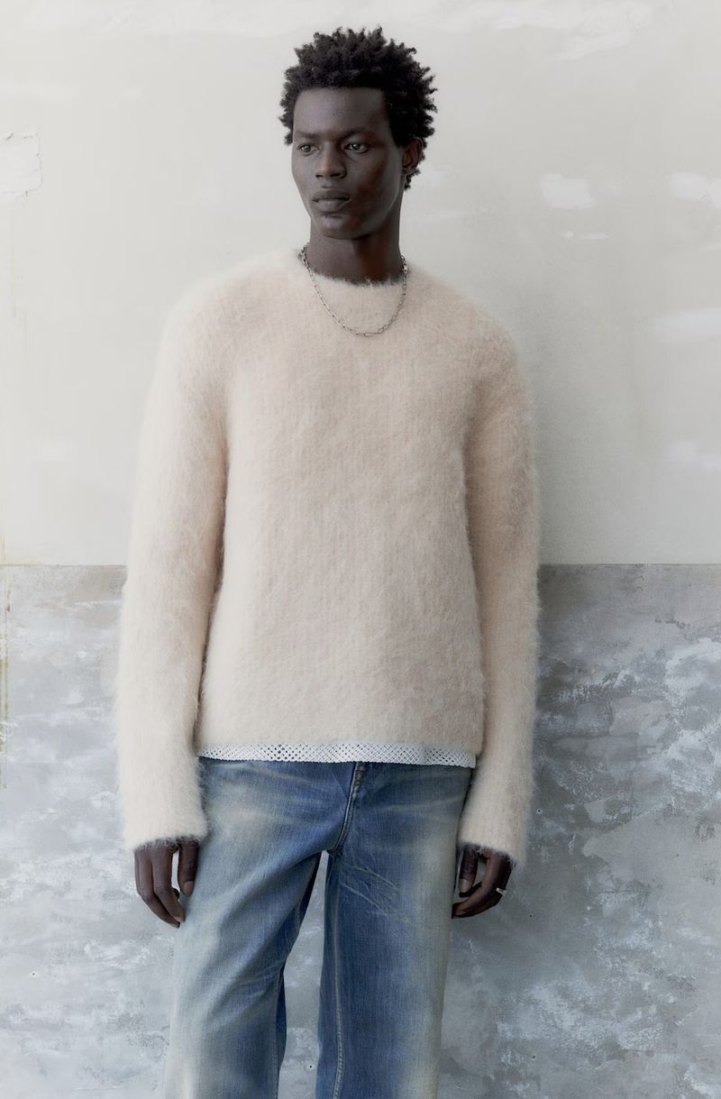 A slender, dark-skinned man in a cream-colored, fuzzy sweater and faded jeans poses against a mottled wall in a medium studio shot.