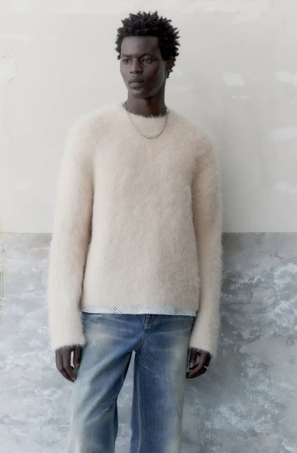 A slender, dark-skinned man in a cream-colored, fuzzy sweater and faded jeans poses against a mottled wall in a medium studio shot.
