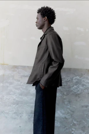 Black male model in profile wearing a dark jacket and trousers against a textured wall background.