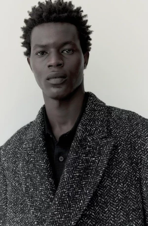 Close-up portrait of a Black man with short dark hair wearing a textured dark herringbone coat against a neutral background.