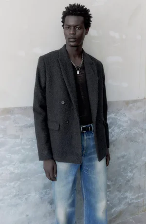 Black man wearing dark grey blazer, black mesh top, and blue jeans against a textured wall