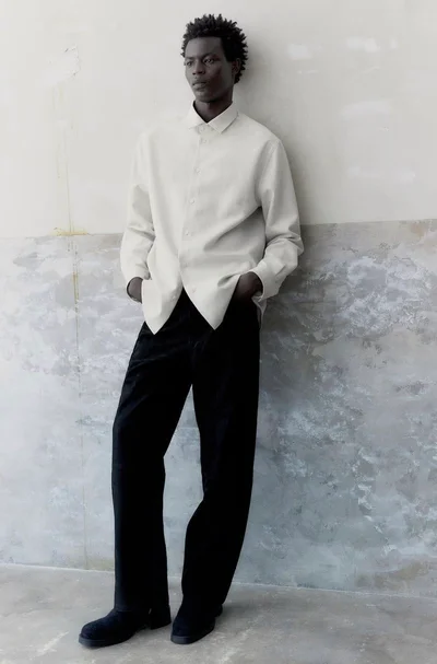Model Mohammed Abubakar leaning against a weathered wall wearing an off-white button-down shirt and black trousers for H&M Men's Atelier.