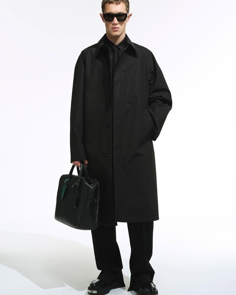 Full-length studio shot of a man in a black trench coat, suit, sunglasses, and black leather bag against a white backdrop.