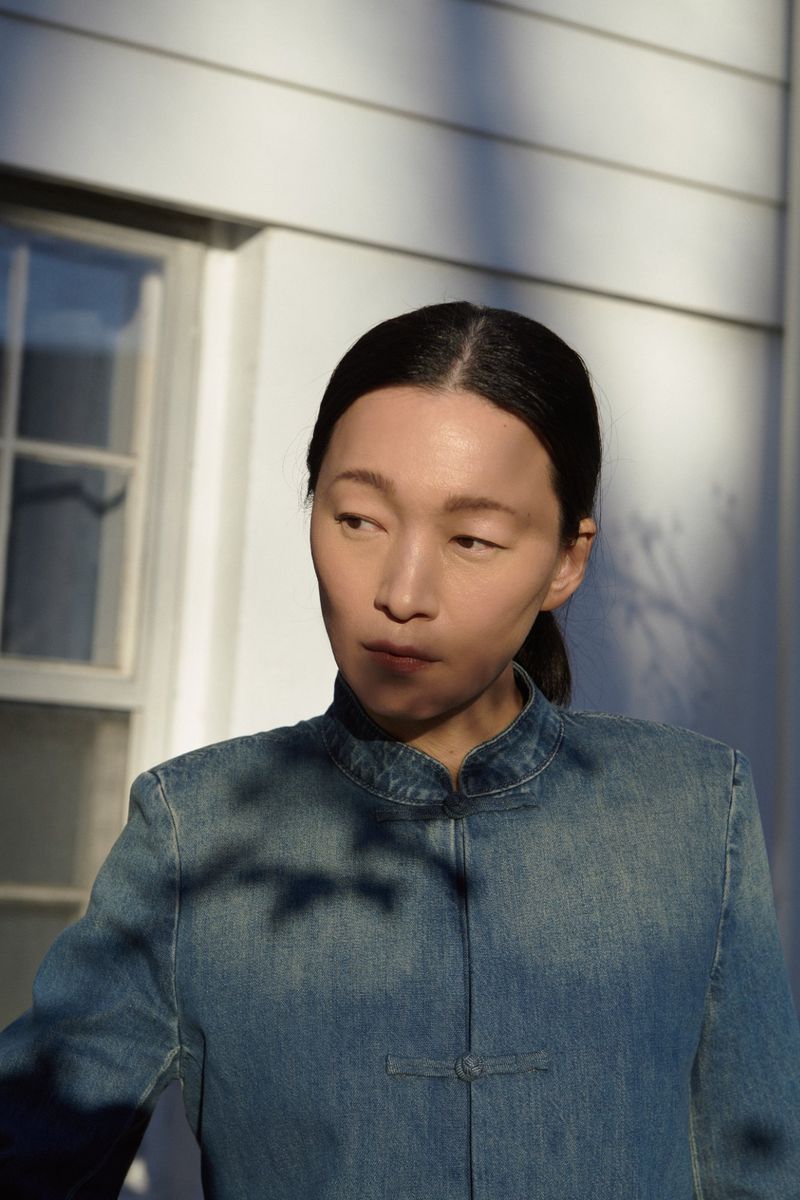 Portrait of an Asian woman in a denim jacket standing outside against a white wall with a window and tree shadows in bright sunlight.