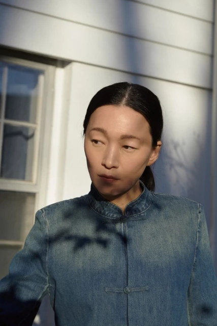 Portrait of an Asian woman in a denim jacket standing outside against a white wall with a window and tree shadows in bright sunlight.