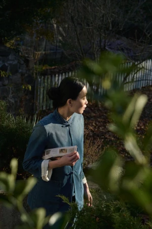 Woman in a denim jacket holding a wrapped object, standing in a garden with green foliage and a stone wall background in soft natural light.