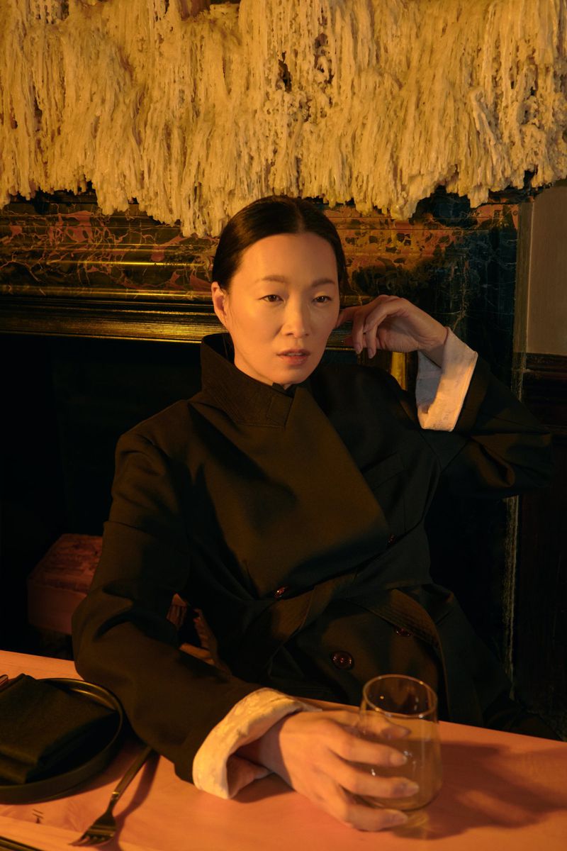 Adult woman in a black jacket sits at a peach-colored table with a glass and tableware in a dimly lit indoor restaurant.