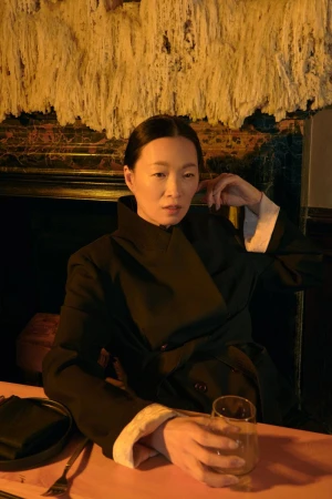 Adult woman in a black jacket sits at a peach-colored table with a glass and tableware in a dimly lit indoor restaurant.