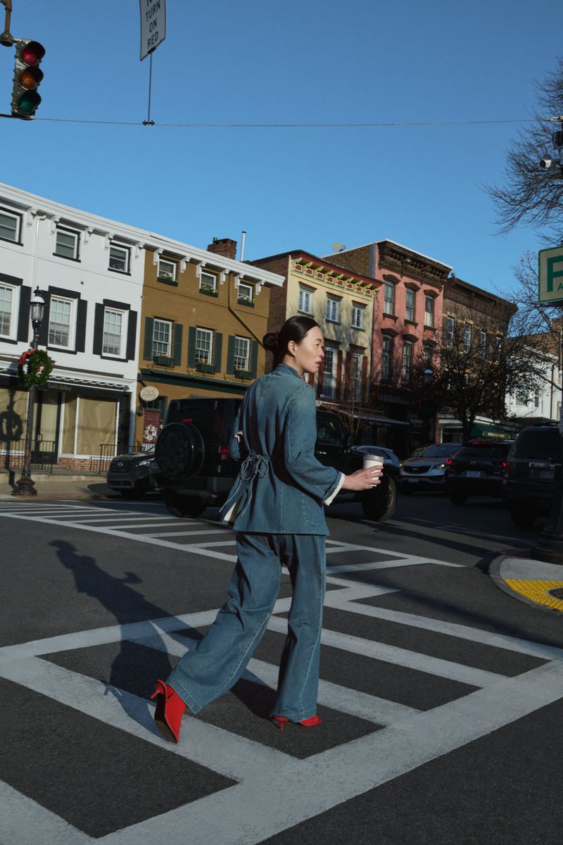 An adult woman in a blue suit walks across a crosswalk in a city, holding a cup of coffee, with buildings and cars visible in the background.