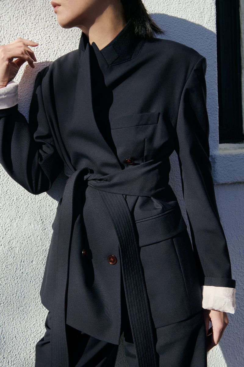 Fashion model in a chic belted blazer and matching pants, posing against an off-white wall in natural light, demonstrating style and elegance.