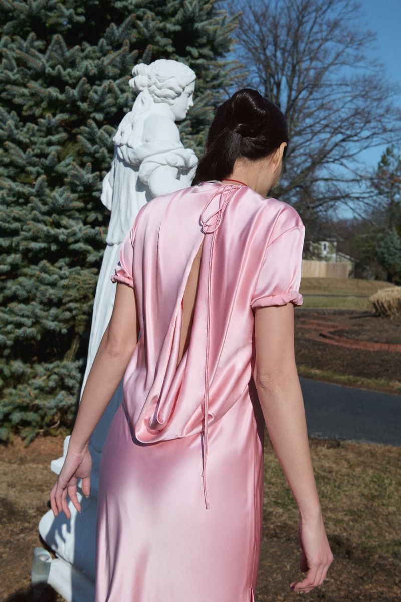 Woman in a pink gown stands near a statue in a garden. The gown has a draped, open back, and the setting includes trees and grass.