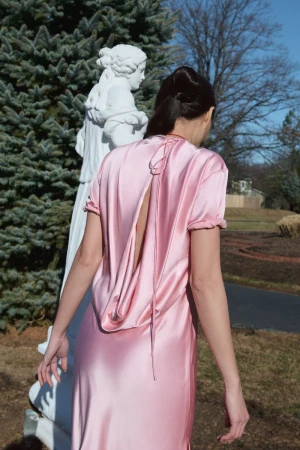 Woman in a pink gown stands near a statue in a garden. The gown has a draped, open back, and the setting includes trees and grass.
