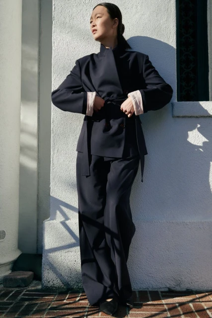 A solitary Asian woman strikes a relaxed pose outdoors, clad in a stylish dark blue suit, illuminated by natural light and subtle shadows.