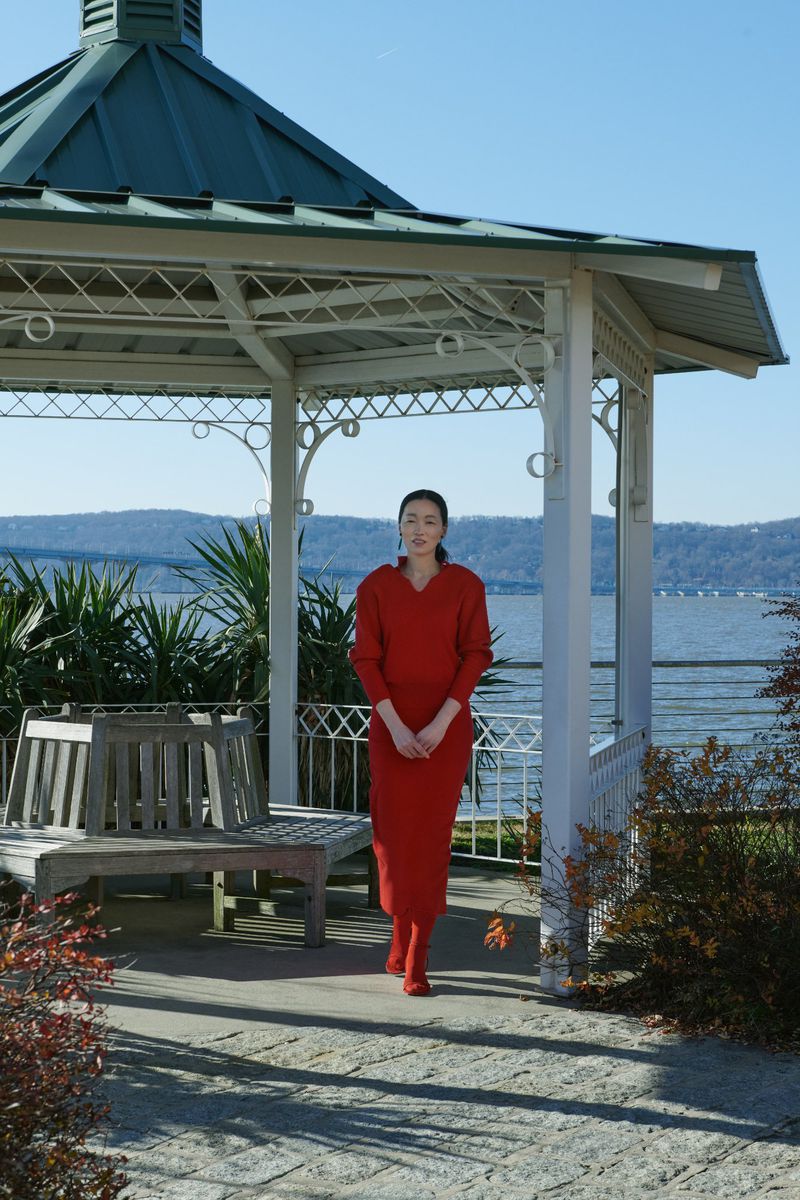 An adult woman stands inside a white gazebo, wearing a red sweater and long red dress, with water and mountains in the background.
