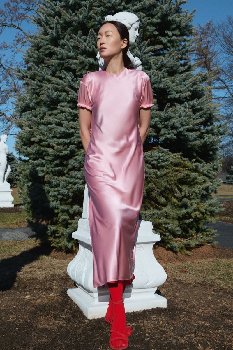 Young woman in a pink dress stands in a park in front of a statue. She is wearing red tights and sandals and poses in natural light.