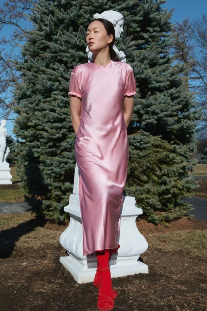 Young woman in a pink dress stands in a park in front of a statue. She is wearing red tights and sandals and poses in natural light.