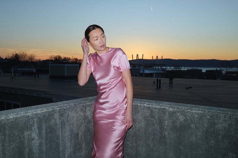 Woman poses on rooftop wearing pink satin dress against sunset backdrop. Fashion photography showcases dress and skyline.