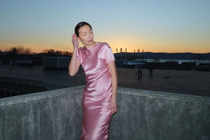 Woman poses on rooftop wearing pink satin dress against sunset backdrop. Fashion photography showcases dress and skyline.