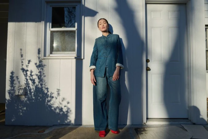Woman in a denim outfit stands outside a white, wood-paneled house, posing near the front door and window, bathed in bright sunlight.
