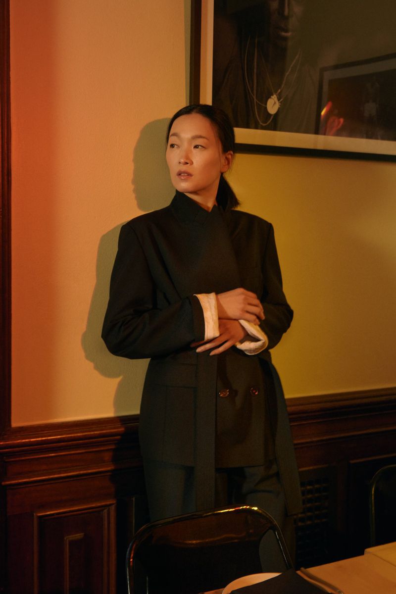 Portrait of Asian woman in elegant dark suit, standing indoors with a serene expression in warm, diffused lighting and simple background.
