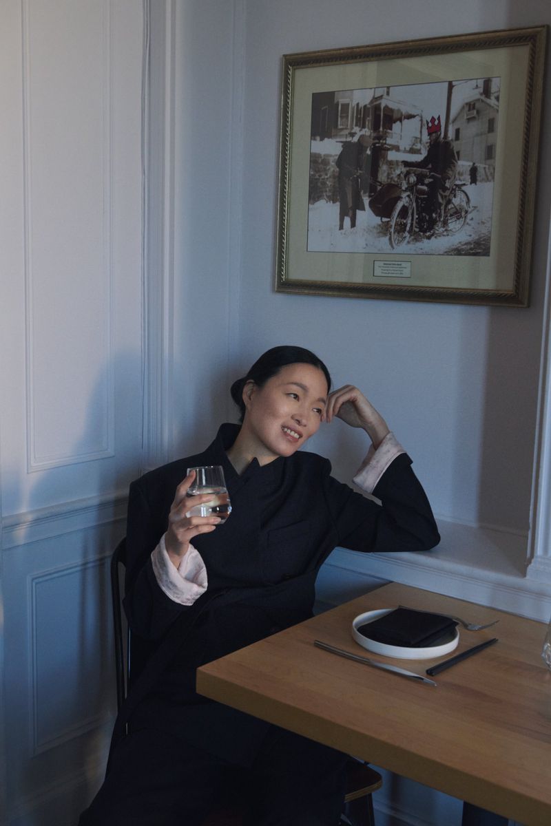 An Asian woman in a black coat seated at a table in a cafe, holds a glass, and smiles near a framed picture on the wall.