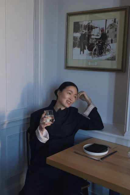 An Asian woman in a black coat seated at a table in a cafe, holds a glass, and smiles near a framed picture on the wall.