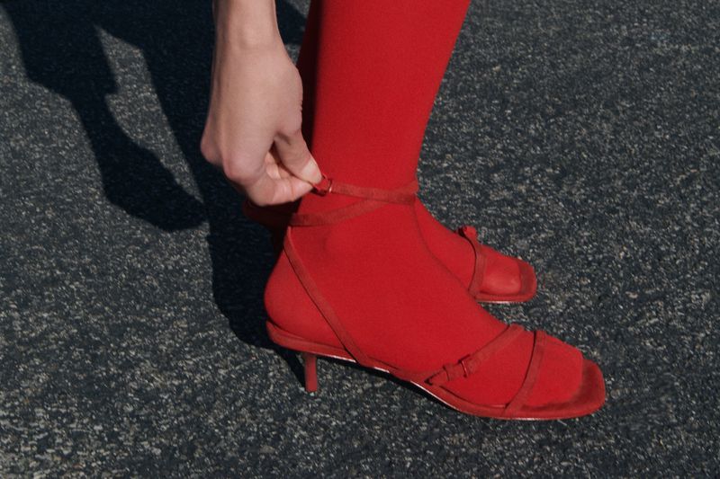 A close-up shot of a woman's leg wearing red tights and strappy heeled sandals, with a hand adjusting the ankle strap outdoors.