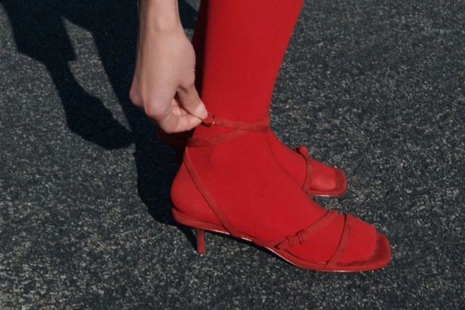 A close-up shot of a woman's leg wearing red tights and strappy heeled sandals, with a hand adjusting the ankle strap outdoors.