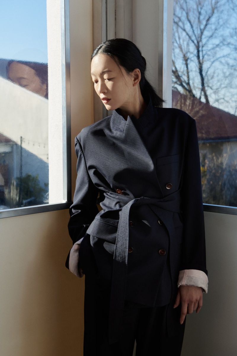 An Asian woman poses indoors near a window, wearing a dark navy blazer and trousers in front of a window with sunlight.
