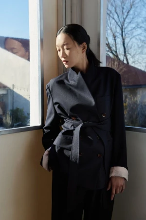 An Asian woman poses indoors near a window, wearing a dark navy blazer and trousers in front of a window with sunlight.