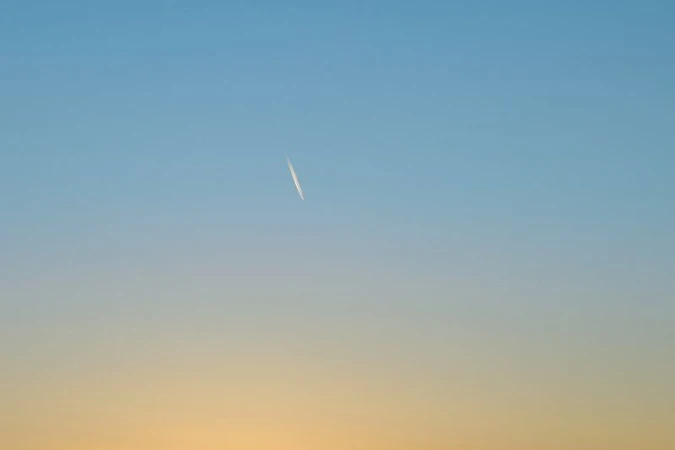 Sky with airplane contrail. Bright daytime scene with wispy vapor trail against a gradient of blue and yellow sky.