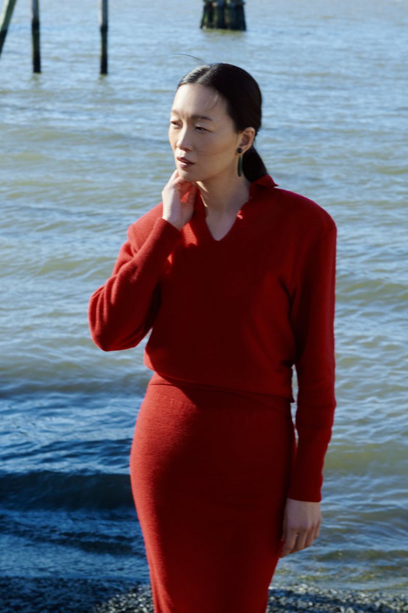 Asian woman in a red dress on a rocky beach by the water, looking off into the distance on a bright day near ocean pilings.
