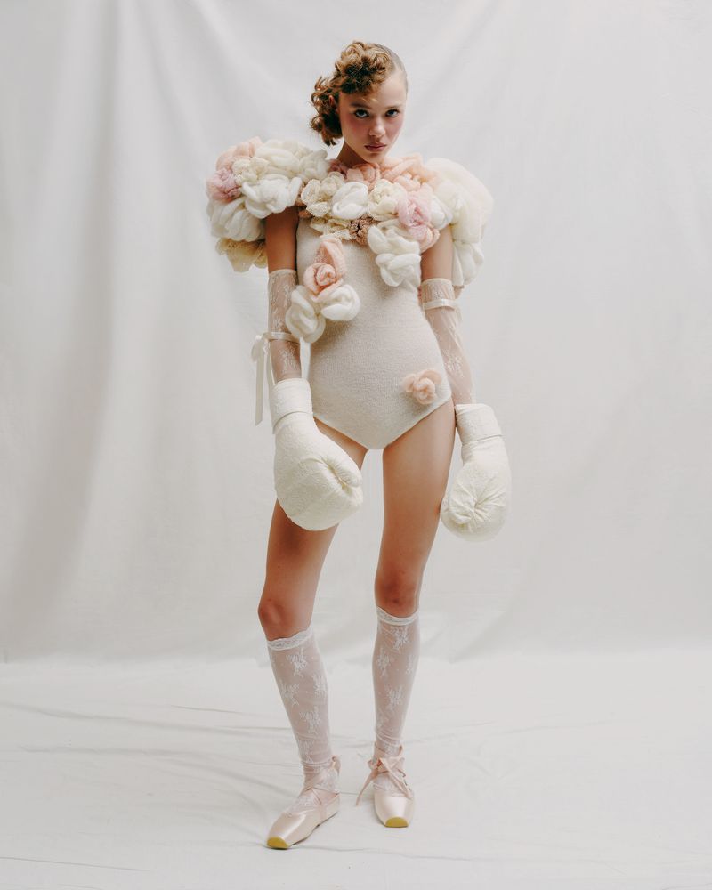 A young woman in a light-colored bodysuit with floral accents poses in a photo studio wearing boxing gloves and pointe shoes.