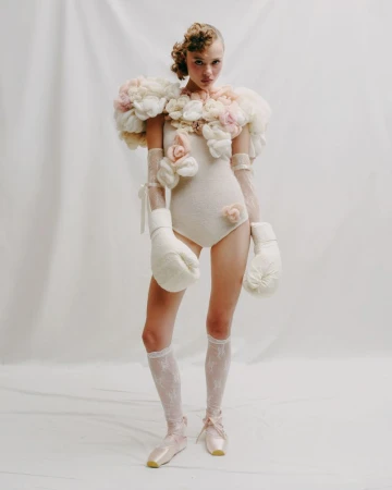 A young woman in a light-colored bodysuit with floral accents poses in a photo studio wearing boxing gloves and pointe shoes.