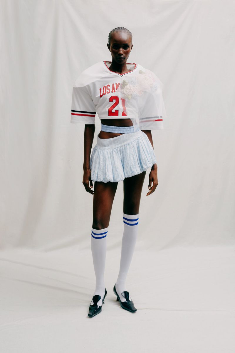 Fashion shot of a Black female model wearing an American football jersey, blue mini skirt, knee-high socks, and black loafers on a white background.