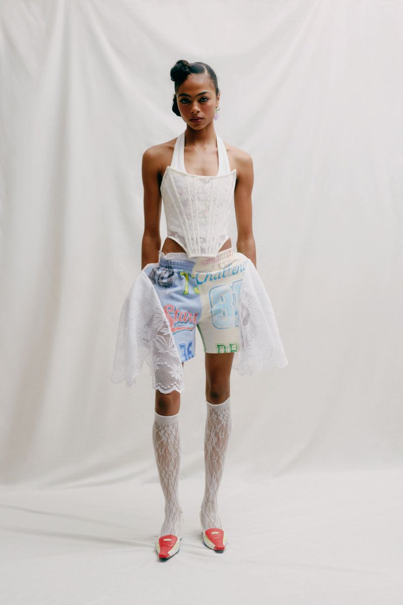 Full shot of a female model posing in a white corset top, shorts, lace socks, and red heels against a neutral backdrop.