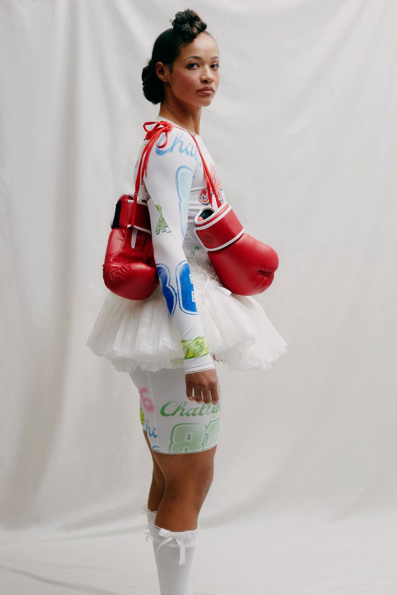 A model in a white dress with colorful text and red boxing gloves posing against a white backdrop in a studio setting.