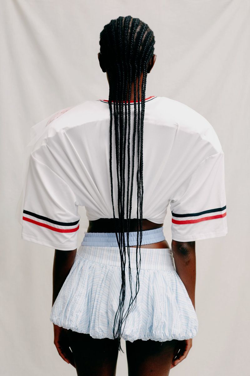 Back view of a woman with braided hair wearing a white cropped t-shirt with red and blue stripes, and a blue textured skirt against white.