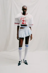 Model wearing a white crop top football jersey and pale blue mini skirt against a white studio background.