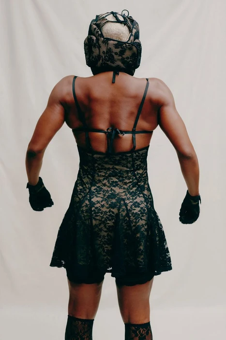 Model from behind wearing a black lace dress and matching headgear against a neutral seamless backdrop.
