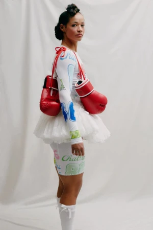 Model wearing a white printed top, tutu skirt, and red boxing gloves against a plain white studio backdrop.