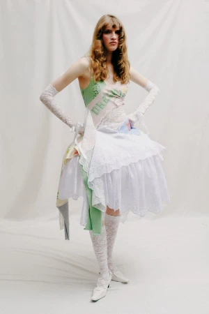 Model wearing a layered white dress and lace gloves standing against a seamless white background for Yuhan Wang SS25.