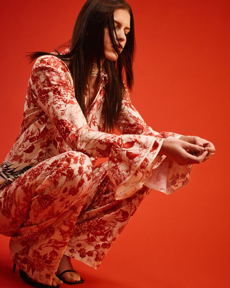 A fashionable adult female is crouching in a photo studio wearing a floral-patterned two-piece outfit with black heeled sandals on a plain red backdrop.