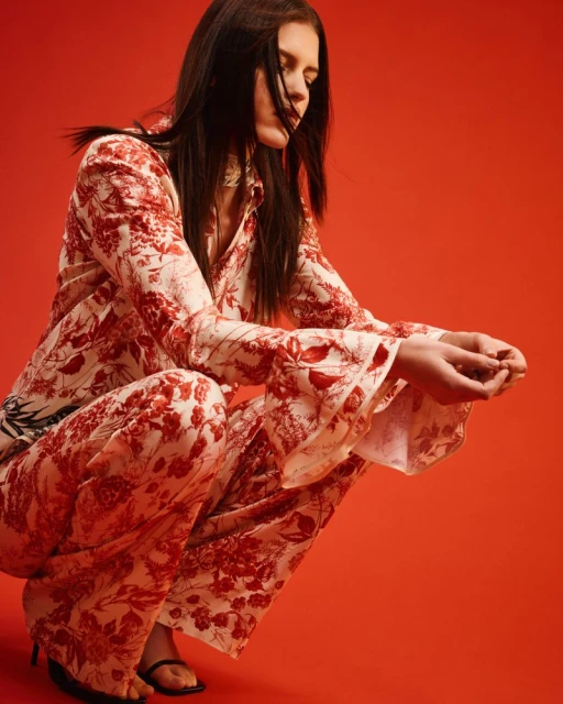 A fashionable adult female is crouching in a photo studio wearing a floral-patterned two-piece outfit with black heeled sandals on a plain red backdrop.