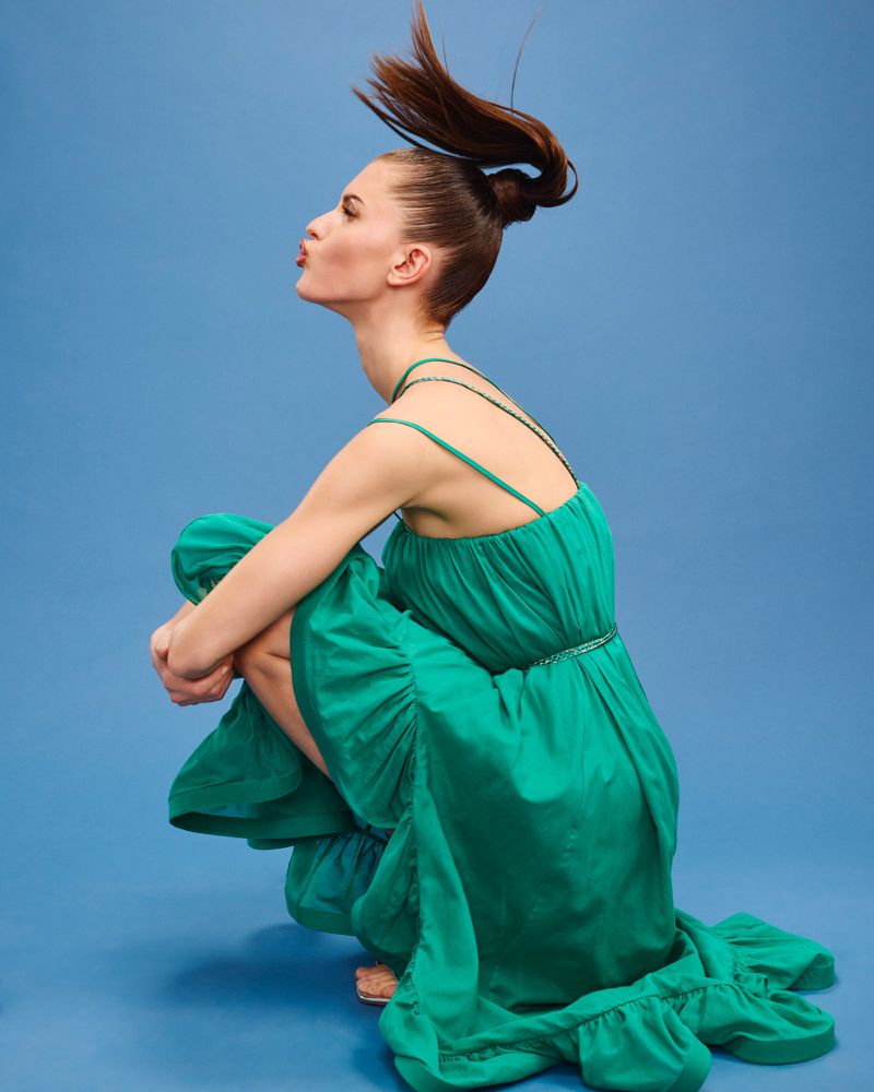 Woman in green dress crouching in a photo studio with a blue background. The subject has her hair up in a high ponytail.