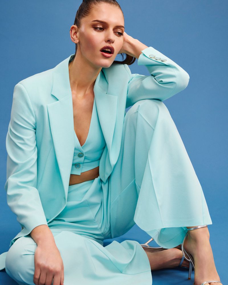 Stylish woman in a light blue blazer, vest, and wide-leg pants poses against a blue background. Silver heels complete the monochrome look.