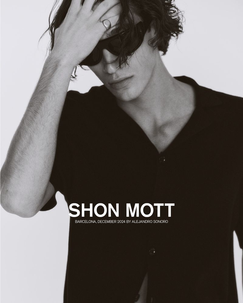 Black and white portrait of a young adult man wearing sunglasses and an unbuttoned shirt, hand on his head, against a light background.