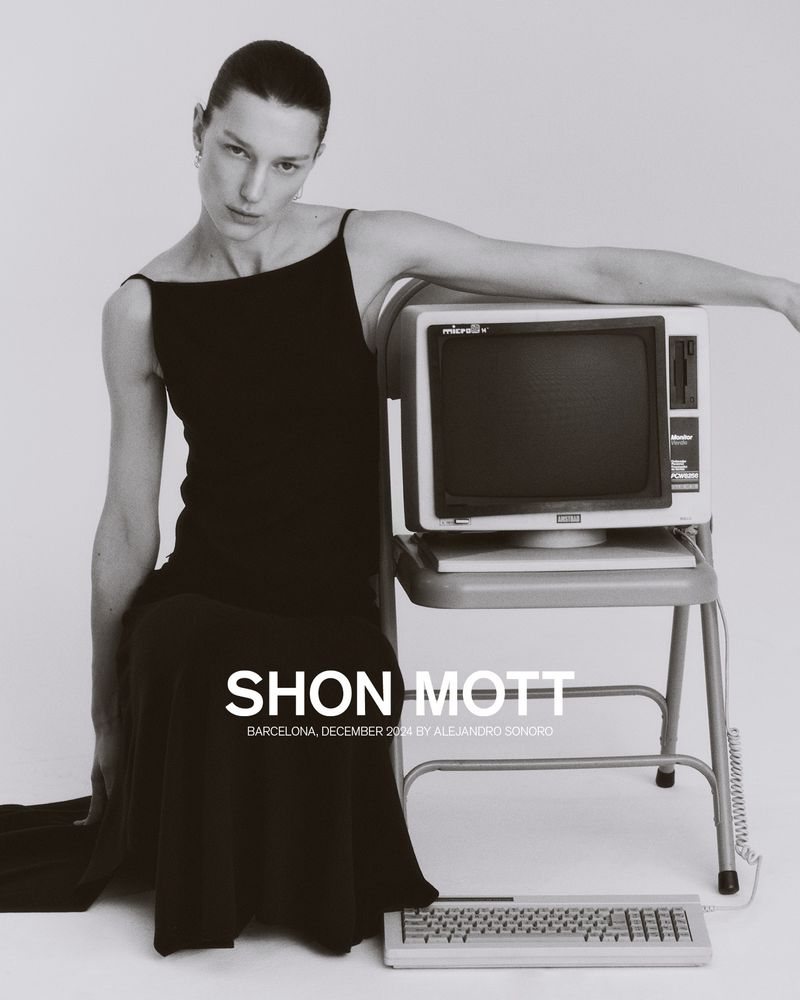 Elegant woman in black dress poses next to vintage computer on grey. Fashion editorial shot; neutral studio backdrop; simple composition.